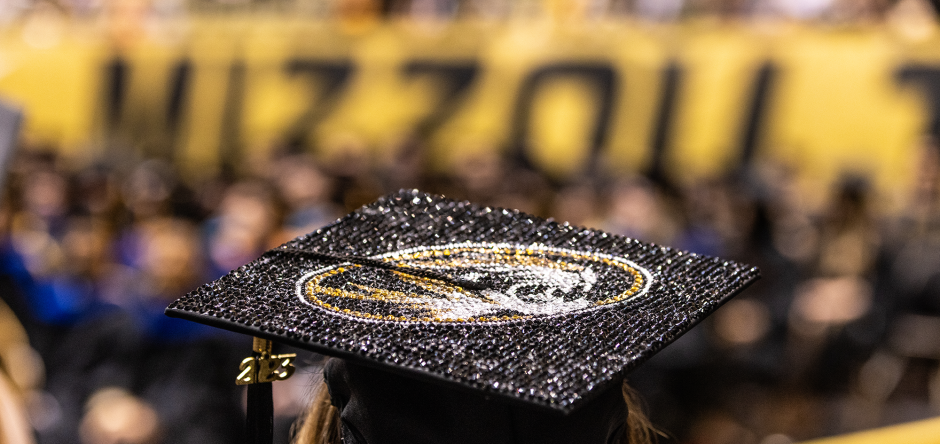 A student at commencement is shown from behind wearing a graduation cap decorated with a Mizzou Tiger.